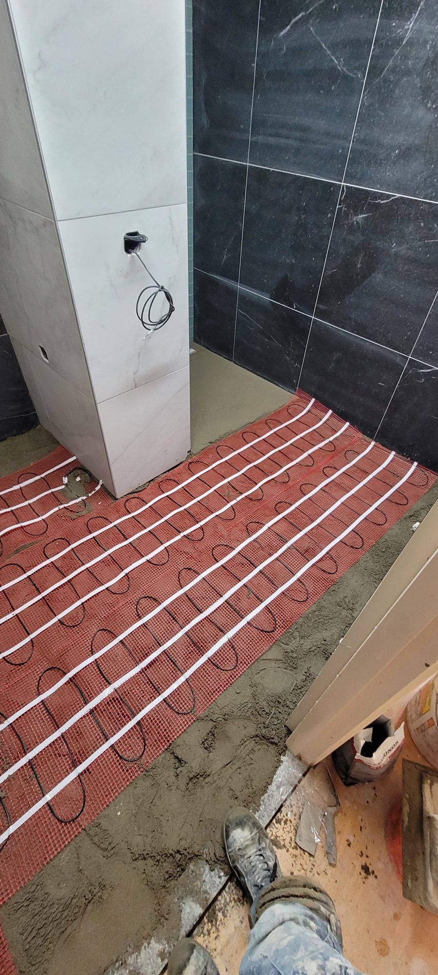Rode matten met elektrische vloerverwarming in de tegellijm, naast zwarte wandtegels in marmerlook
